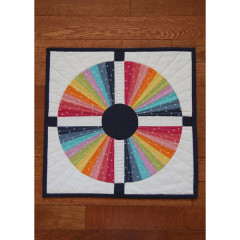Draufsicht auf den fertigen Mini-Quilt mit vierteiligem Regenbogen-Kreismuster und dunkelblauem Kreuzrahmen auf Holzboden.